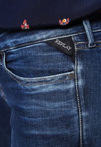 Jeans de denim azul oscuro con un acabado desgastado, diseño de cinco bolsillos y una etiqueta tejida negra con "REPLAY" cerca de la cinturilla.