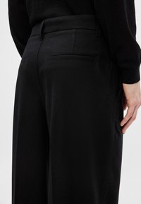 Calças pretas com um corte ajustado, apresentando dois bolsos traseiros, uma textura suave e um brilho subtil. Projetadas para uma aparência elegante.