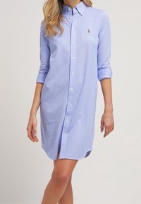 Helles blaues Hemdkleid mit langen Ärmeln, die an den Bündchen aufgerollt sind, mit einem kleinen Logo auf der Brust und einem geraden Saum.