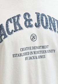 Hvid bomulds-T-shirt med fedt mørkeblåt tekst "JACK & JONES" og yderligere brandingdetaljer nedenunder i en mindre skrifttype.