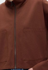 Brown zip-up jacket with two front pockets, worn over blue jeans, showing close-up of fabric texture and zipper detail.