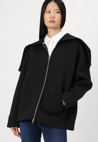 Hoodie oversize noir avec une grande capuche, fermeture éclair, poches avant et un logo "EXT" texturé, fabriqué en tissu doux.