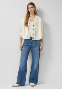 Kremowy sweter kardigan z dzianiny na napy, noszony na jasnym topie na ramiączkach, zestawiony z jasnoniebieskimi szerokimi dżinsami i beżowymi trampkami.