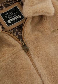 Giacca in pile beige con superficie tessurizzata, dotata di cerniera bronzo e etichetta del marchio. La fodera interna presenta un motivo geometrico in tonalità di marrone.
