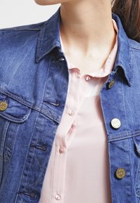 Denim jacket with a classic collar, two chest pockets, and gold-tone Lee buttons. Underneath, a light pink blouse with a soft texture.