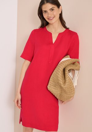 Kvinde smilende iført en rød knælængde kjole med korte ærmer, holder en sammenfoldet vævet stråtaske mod en beige baggrund.