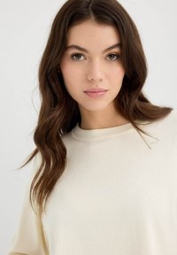 Suéter beige claro con un escote redondeado, hecho de una tela suave, con mangas largas y un corte relajado, mostrado en un modelo con cabello ondulado.