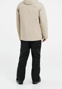 Veste beige isolante avec capuche et pantalon de neige noir. La veste présente une texture lisse et un logo, le pantalon est équipé de poches zippées et d'une coupe décontractée.