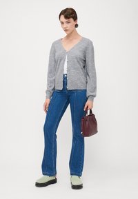 Grey knit cardigan with round neckline, pearl buttons, paired with high-waisted blue flared jeans and green slip-on shoes, holding a burgundy bag.