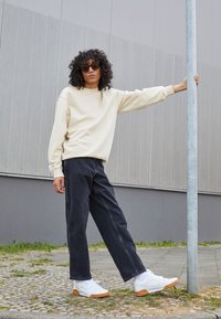 Felpa beige chiaro, pantaloni di jeans scuri, sneakers bianche con suole color gomma e occhiali da sole marroni. Il soggetto è appoggiato a un palo all'aperto.