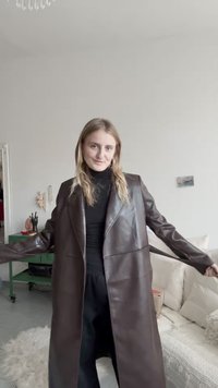 Cămașă lungă din piele maro cu un design structurat, rever în formă de V și buzunare laterale, purtată peste un pulover negru cu guler și pantaloni largi.