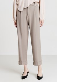 Pantaloni beige su misura con una vestibilità rilassata, dotati di orli cuffi e una superficie liscia, abbinati a décolleté nere a punta.
