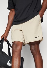 Beige athletic shorts with a black Nike logo, made from a lightweight, textured material. Pairing with a black sports bag held in hand.