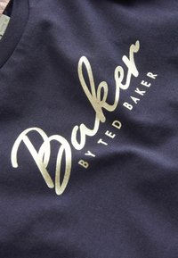 Tumši zils audums ar spīdīgu zelta "Baker by Ted Baker" logotipu, kas diagonāli izdrukāts uz virsmas.
