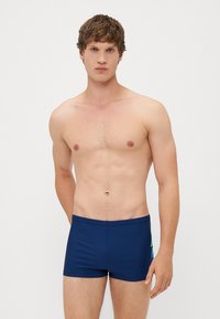 Marinblå badshorts med en slät textur, med en minimalistisk design och subtila geometriska detaljer på sidan.