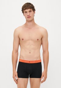 Schwarze Herren-Boxershorts mit einem orangefarbenen Tommy Hilfiger-Logo-Bund. Der Stoff wirkt glatt und eng anliegend, wobei die Körperform betont wird.