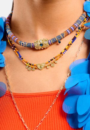 Trois colliers superposés sur un cou de couleur chair. Le supérieur est composé de perles multicolores avec un fermoir décoratif doré, le inférieur présente un pendentif en forme de cœur et des accents dorés.