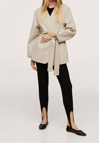 Beige wikkelvest met een ceintuur, lange mouwen, gecombineerd met zwarte leggings met openingen bij de enkels en zwarte balletschoenen.