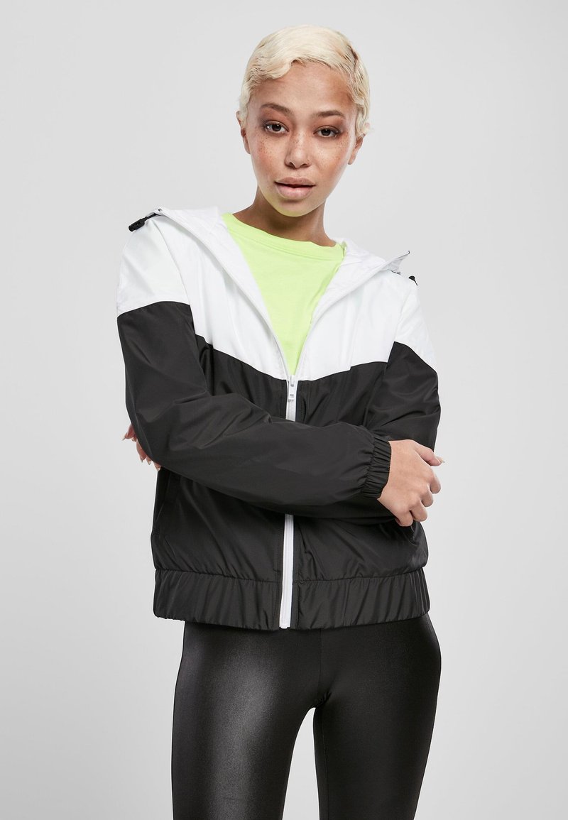 Svartvitt zip-up-jacka med huva, utrustad med elastiska ärmslut och dragsko i fållen, bärs över en ljusgrön tröja och svarta leggings.