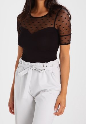 Kvinde iført en sort, gennemsigtig kortærmet bluse med polkaprikker, kombineret med højtaljede, lysgrå paperbag-bukser, der er bundet med et bælte i taljen.