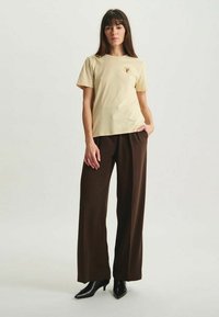 T-shirt en coton jaune clair à manches courtes avec un petit logo rond sur la poitrine, associée à un pantalon large marron et des chaussures à talons noires.