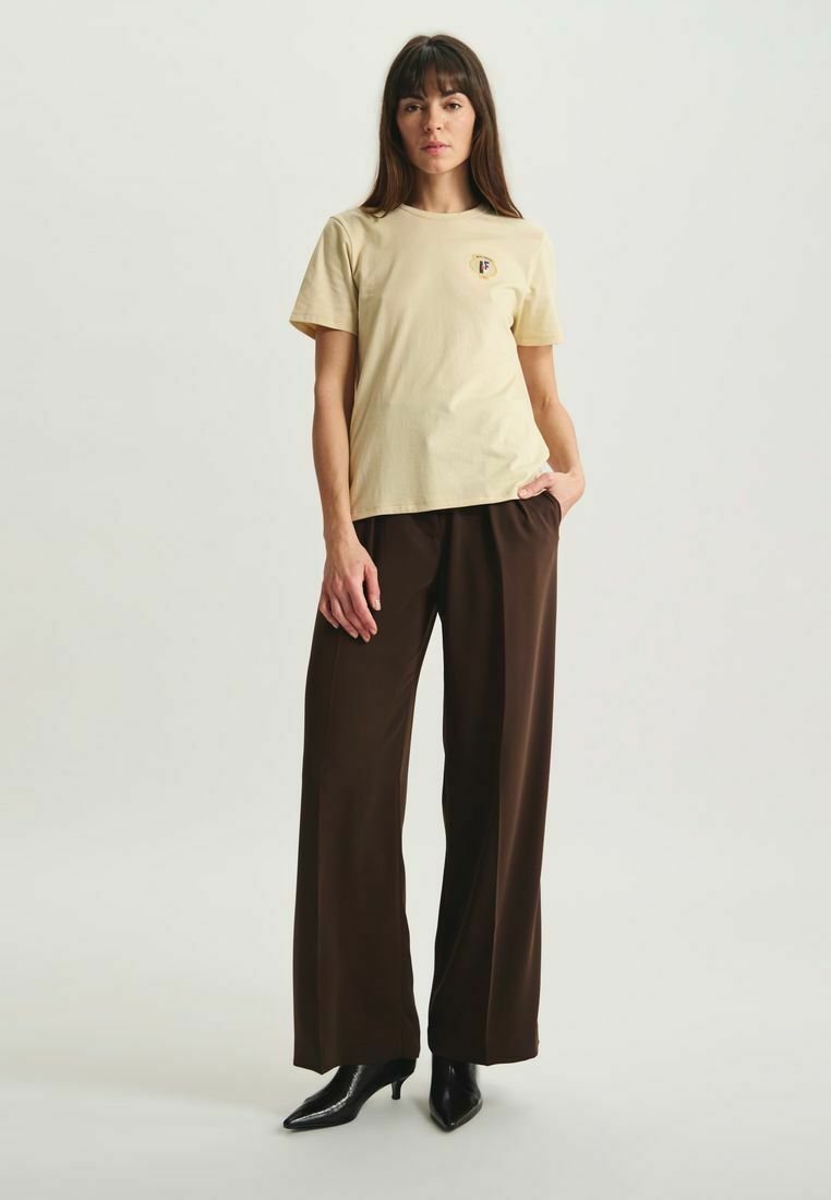 T-shirt en coton jaune clair à manches courtes avec un petit logo rond sur la poitrine, associée à un pantalon large marron et des chaussures à talons noires.