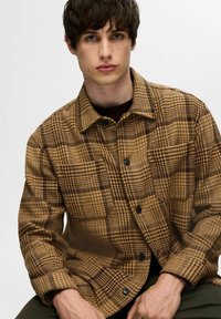 Brązowa i żółta koszula w kratę z kurtką ma zapięcie na guziki, duży kołnierz oraz dwie kieszenie na piersi. Teksturowany materiał dodaje głębi.