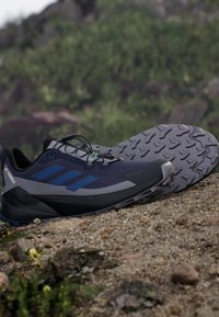 Scarpa sportiva nera e blu scuro con suola robusta, parte superiore in rete, lacci e logo; progettata per l'uso all'aperto con presa testurizzata.
