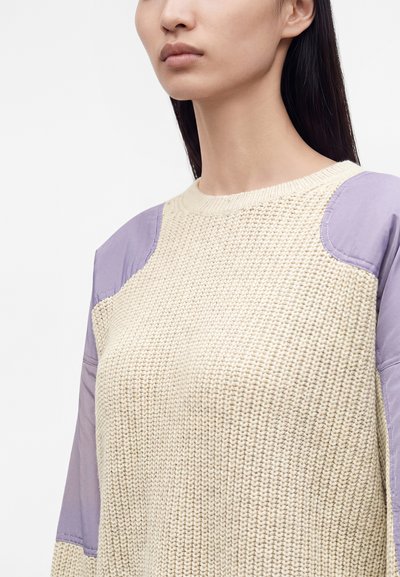 Femme portant un pull en maille crème avec des patchs en tissu violet clair sur les épaules et les manches, visible des épaules au menton.
