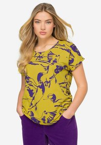 Top amarillo de manga corta con un patrón de pájaros morados, que presenta un cuello redondo y un corte holgado, acompañado de pantalones de pana morados.