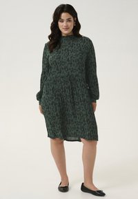 Robe verte avec un motif animal noir, col haut et manches longues. La taille est froncée, créant une coupe ample. Des ballerines noires complètent le look.