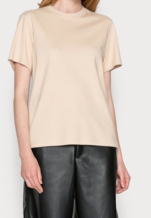 Person trägt ein beiges kurzärmeliges Oberteil mit Rundhalsausschnitt und schwarze Kunstlederhosen, steht vor einem einfarbigen Hintergrund.