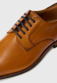 Zapato de vestir de cuero marrón con acabado suave, seis cordones negros y detalles de costura sutiles. Diseño de punta redonda y suela plana.