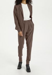 Brązowa marynarka w zestawie z pasującymi spodniami z elastycznym pasem, połączona z białą bluzką. Gładki materiał, minimalistyczny design i eleganckie botki za kostkę.