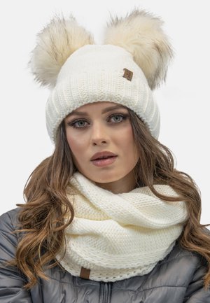 Hvid strikket beanie med to faux fur pom-poms, ledsaget af et hvidt strikket infinity-scarf med et struktureret mønster.