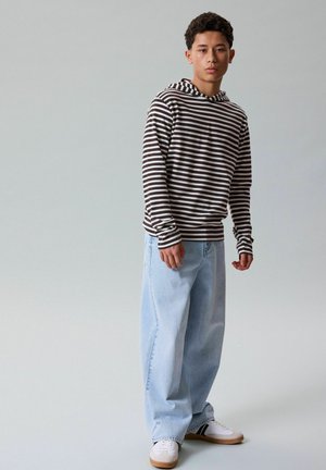 Ung mand står iført sort-hvid stribet hættetrøje, vide lyseblå jeans og hvide sneakers med sorte striber på ensfarvet baggrund.