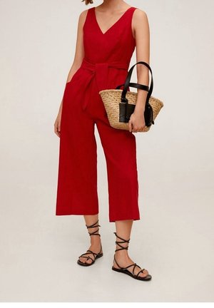 Czerwony kombinezon bez rękawów z dekoltem w serek, wiązaniem w talii i szerokimi, krótkimi nogawkami, zestawiony ze słomkową torebką i czarnymi sandałami.