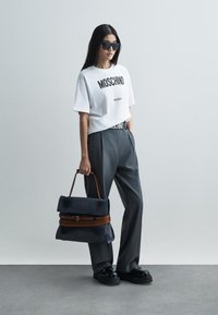 Camiseta de algodão branca com texto preto "Moschino Milano", calças cinzentas de cintura alta, sapatos pretos robustos e uma mala azul-marinho com detalhes castanhos.