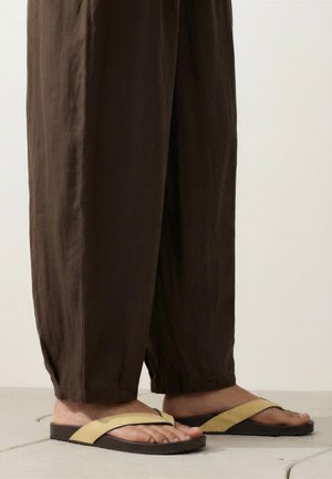Picioare purtând sandale plate bej și negre asortate cu pantaloni largi maro închis, stând pe podea placată cu gresie.