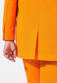 Oranssi puku takki, jossa on halkio, kapea muotoilu ja napitusyksityiskohta hihassa. Silkkinen pinta ja kirkas, tasainen väri. Käsi näkyvissä.