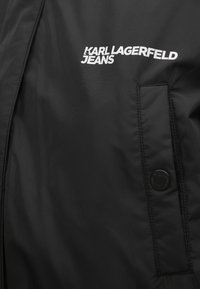 Čierna tkanina s našitým vreckom a čiernym patentným gombíkom, s bielym logom "KARL LAGERFELD JEANS" na ľavej hornej strane.