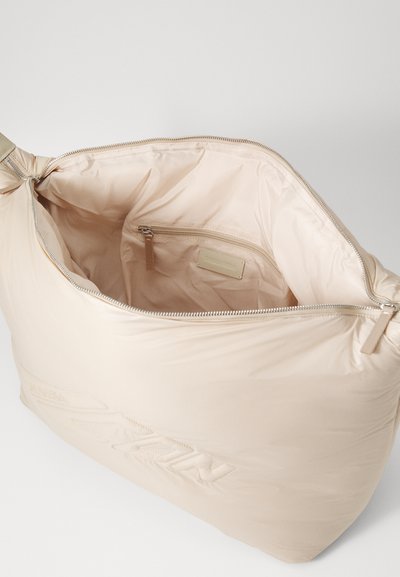 Beige padded bag with a zip closure and inner pocket. Smooth texture, rounded shape, and embossed logo detail on one side.