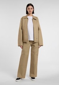 Beige zip-jacka med spetsig krage, i kombination med matchande vida byxor, som har fickor och en logotyp. Vit T-shirt under.