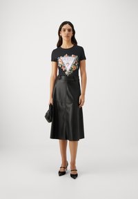 T-shirt noire avec un motif floral et le texte "GUESS", associée à une jupe midi en cuir noire, des chaussures à talons et un petit sac noir.