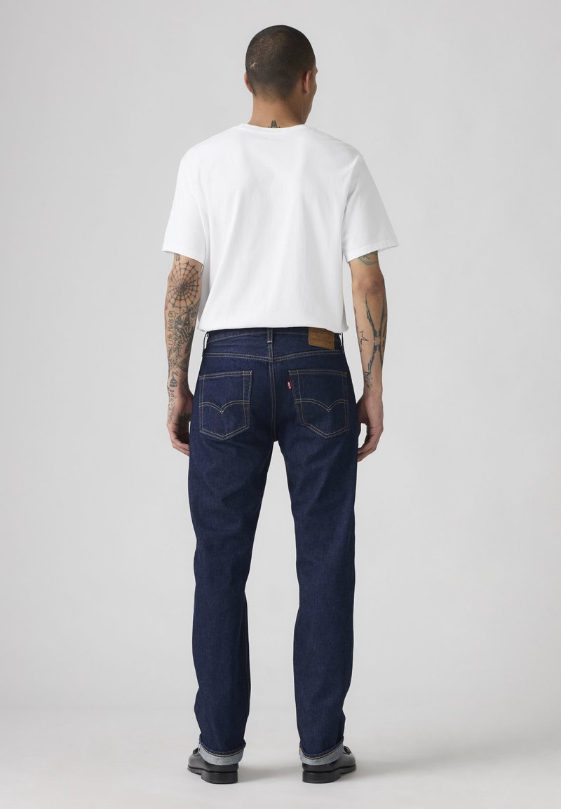 Hombre con tatuajes que lleva una camiseta blanca y vaqueros azul oscuro, de pie con la espalda hacia la cámara frente a un fondo liso.