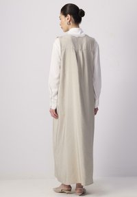 Dlhé, bezrukávové šaty vo svetlej béžovej farbe, s košeľou s gombíkmi pod nimi. Hladká textúra s jemným drapovaním, doplnené slingback topánkami.