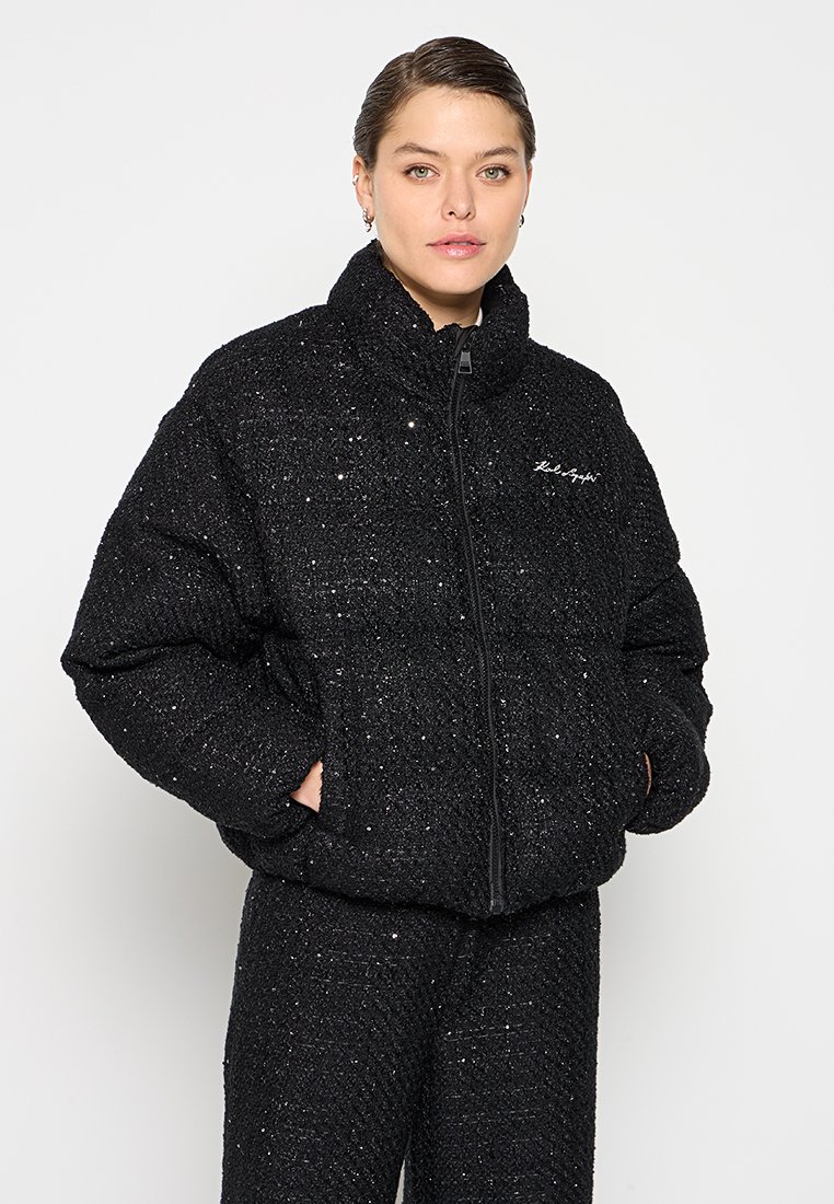 Schwarze Pufferjacke aus einem strukturierten Stoff mit leichtem Glanz, mit Reißverschluss und hohem Kragen. Besticktes Logo auf der Brust.