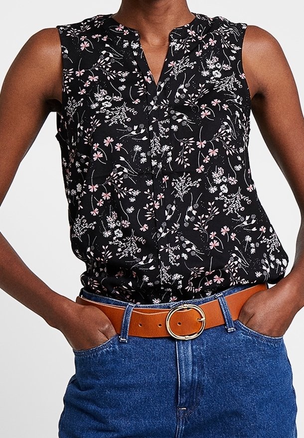 Blouse noire sans manches avec motif floral blanc et rose, rentrée dans un jean bleu avec une ceinture marron avec une boucle ronde dorée.