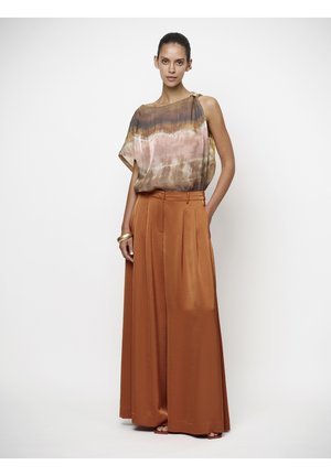 Vrouw draagt een mouwloze, meerkleurige top en wijd uitlopende, hooggetailleerde broek in verbrand oranje, staand tegen een effen witte achtergrond.