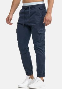 Pantalones cargo de color navy con puños elásticos, cinturilla con cordón, múltiples bolsillos y una textura suave. Combinados con zapatillas blancas.